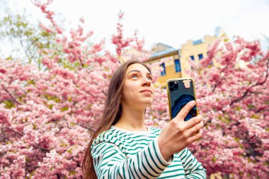 Akıllı telefon özçekim portresi için poz veren genç bir bayan. Kafkasyalı bir kadın kiraz çiçeği sakura çiçeklerinden selfie çekiyor. Bahar selfie 'si, yaşam tarzı.