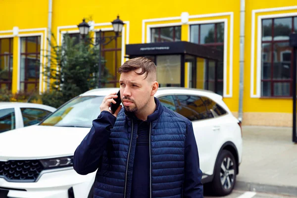 Frowning tense sullen or sad concentrated young caucasian 34 years old man with goatee beard talks on smartphone on street near white car. Waist up lifestyle business portrait, difficult conversation