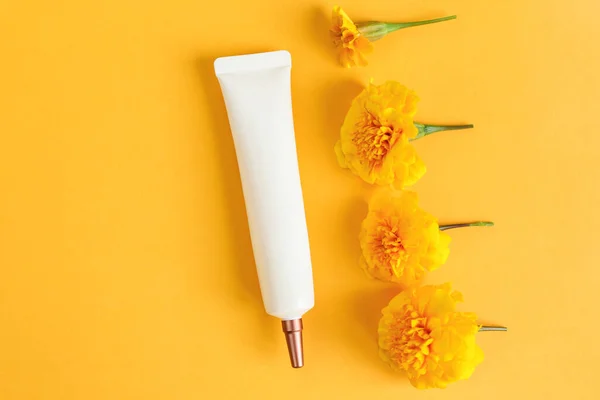 Face and eye cream squeeze cosmetic white tube with long nozzle and bronze screw cap and yellow marigold flowers line on yellow background. Skincare, moisturizer cream, mockup. Front view.