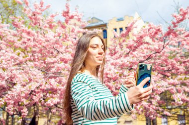 Portrait making selfie female on smartphone on cherry blossom sakura tree. Young smiling caucasian woman is taking selfie portrait with mobile phone. Summer Spring waist up lifestyle portrait. clipart