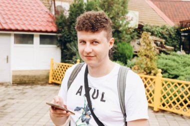 Beyaz, kıvırcık saçlı, akıllı telefon tutan, gülümseyen ve kameraya bakan genç bir adam. Portre.