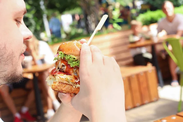 Kafkasyalı genç bir adam kaldırımdaki kafede hamburger ya da sığır burger yiyor, güneşli bir günde brunch yapıyor. Yakın çekim, fast food, portre