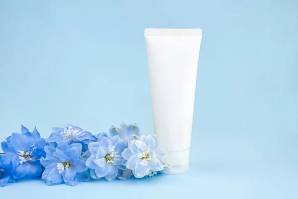 Mockup white squeeze bottle plastic tube for branding of medicine or cosmetics - cream, gel, skincare, moisturizer. Cosmetic bottle container and blue Delphinium flower on blue background. Front view