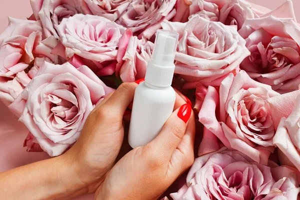 White cosmetic small spray bottle in female woman arms hands on pink rose flowers background, mockup. Antibacterial liquid, natural organic oil, body mist, sanitizer, perfume, sunscreen, deodorant