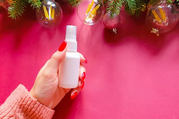 White cosmetic spray bottle in female hand with red nails, fir tree, retro lamps on red background. Antibacterial liquid, natural organic oil, body mist, sanitizer, perfume, sunscreen, deodorant