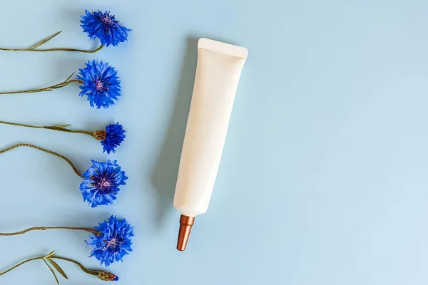 Face and eye cream squeeze cosmetic tube with long nozzle and bronze screw cap and knapweed cornflower flowers line border on blue background. Moisturizer, skincare, makeup, moisturizing cream. Mockup