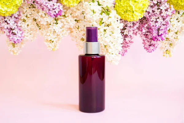 Brown cosmetic spray bottle,  purple and white lilac and green viburnum flowers on pink background. Mockup. Skincare beauty, liquid antibacterial spray, body oil, natural body mist. Front view