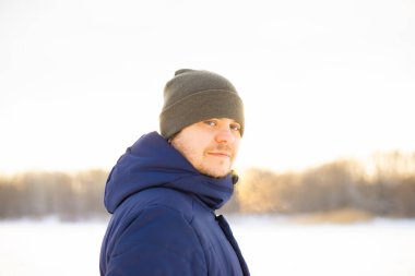 Young man in winter on a snowy lake, portrait. European caucasian white male in hat and down jacket in sun rays.