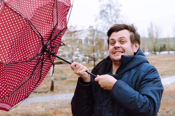 Shocked European white young male man fool around with umbrella in city park, strong storm wind, rain and bad weather. Wind broke umbrella. Autumn waist up lifestyle portrait.