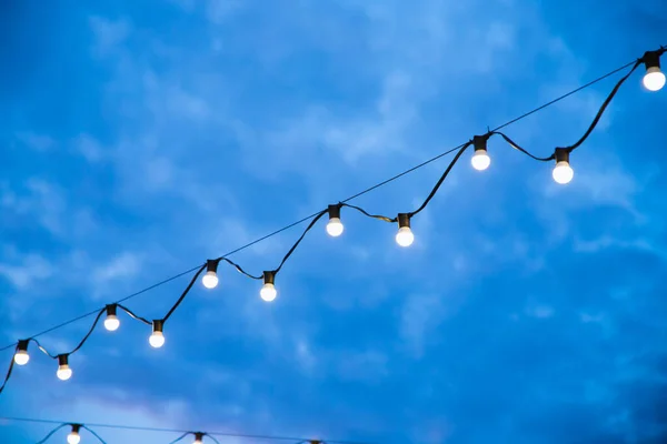 Street outside lighted retro light bulbs garland on night sky. String of light bulb decoration for outdoor activities, party, concert, festival, fun fair.