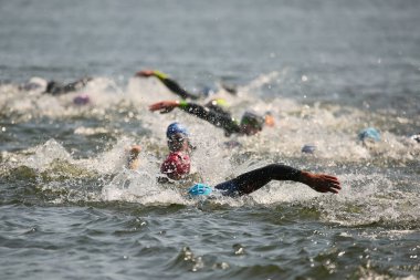 Triatlon sporcuları grup olarak açık denizde yüzerler, dalgıç kıyafeti ve şapkalar giyerler, yarışma sırasında su sıçratırlar.