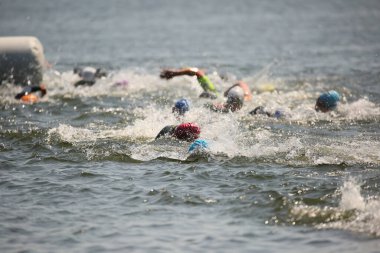Triatlon sporcuları grup olarak açık denizde yüzerler, dalgıç kıyafeti ve şapkalar giyerler, yarışma sırasında su sıçratırlar.