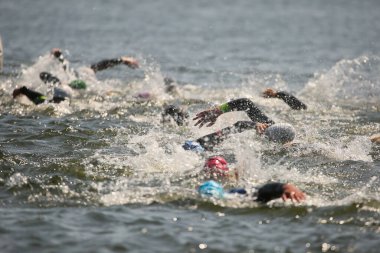 Triatlon sporcuları grup olarak açık denizde yüzerler, dalgıç kıyafeti ve şapkalar giyerler, yarışma sırasında su sıçratırlar.
