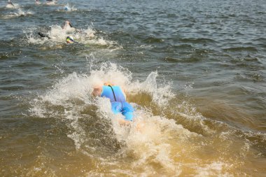 Triatlon sporcuları grup olarak açık denizde yüzerler, dalgıç kıyafeti ve şapkalar giyerler, yarışma sırasında su sıçratırlar.
