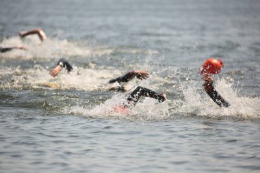Triatlon sporcuları grup olarak açık denizde yüzerler, dalgıç kıyafeti ve şapkalar giyerler, yarışma sırasında su sıçratırlar.