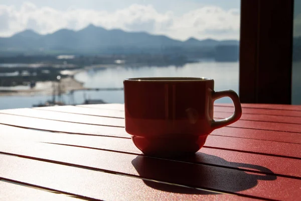 A cup of coffee on a red table. View of the lake, sea, ocean, river. Shadows, backlight