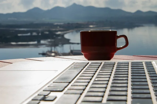 Cup with coffee on a red table.Laptop, keyboard close-up. View of the lake, sea, ocean, river. Shadows, backlight