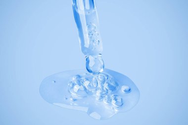 Şeffaf kozmetik jel, baloncuklu büyük bir kabarcığın içinde göz damlasından doğrudan arka plana akar. Mavi arka planda. Hyaluronik asit, tonik, tonik, jöle, krem. Kozmetik.