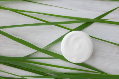 Uzun yeşil yapraklarla çevrili beyaz ahşap arka planda beyaz kozmetik krem.