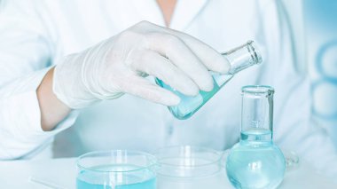 Laboratuvar ayarları: Asistan dikkatle şişeleri temiz mavi sıvıyla doldurur. Eldivenli eller. Beyaz laboratuvar önlüğü. Cam ekipman..