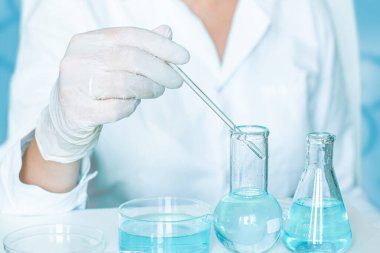 Laboratuvarda, laboratuvar asistanı manipülasyonlar yapar. Eldivenler ve beyaz bir bornoz giymişti. Bardak kullanılmış. Sıvı açık mavi.