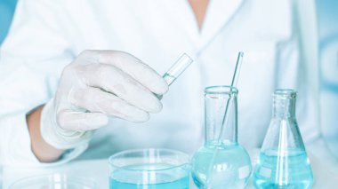 Laboratuvar ortamı: Yardımcı laboratuarda eylemleri gerçekleştirir. Eldivenler ve beyaz bir bornoz. Cam eşyalar ve berrak mavi sıvı kullanılmış..