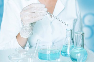 Laboratuvarda asistan materyalleri manipüle ediyor. Eldivenleri ve beyaz giysileri vardı. Cam eşyalar kullanılıyor. Kullanılan şeffaf mavi sıvı.