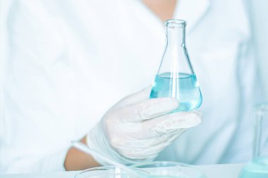 Laboratuvarda, laboratuvar asistanı mataranın sıvısı üzerinde işlem yapıyor. Eldivenler, beyaz bornoz. Cam enstrümanlar kullanılmış. Açık mavi sıvı mevcut.