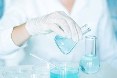 Bilimsel kurulum: Asistan mataranın sıvısıyla manipülasyon yapıyor. Eldiven giyip, beyaz bir bornoz giyip, cam aletlere dokunuyor. Açık mavi sıvı mevcut.