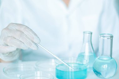 Laboratuvarda yardımcı, mataranın sıvısıyla manipülasyon yapıyor. Eldivenler, beyaz bornoz ve kullanılan cam enstrümanlar. Açık mavi sıvı içeriyor.