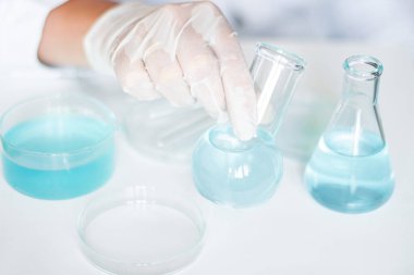 Laboratuvar asistanı mataradaki sıvıyı manipüle ediyor. Eldiven giyiyor, beyaz bornoz giyiyor, cam eşyalar kullanıyor. Açık mavi sıvı içeriyor.