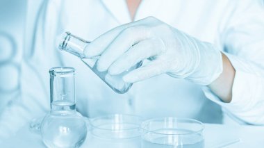 Bilimsel sahne: Asistan laboratuvar ortamında manipülasyonlar yapıyor. Laboratuvar önlüğü. Cam aletler kullanılmış. Mavi bir labaratuvarda. Şeffaf sıvı ile araştırma.