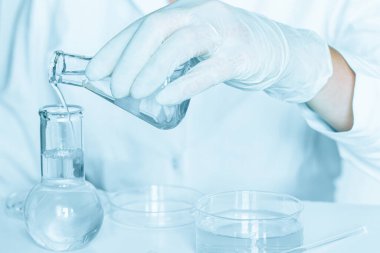 Laboratuvar kurulumu: Asistan laboratuvar koşullarında manipülasyon yapar. Laboratuvar önlüğü giyiyorum. Cam aletler yerleştirildi. Mavi laboratuvar içeriği. Şeffaf sıvıyla araştırma yapıyor..