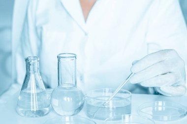 Bilimsel ayar: Laboratuvar asistanı laboratuvar ortamında manipülasyonlar yapıyor. Laboratuvar önlüğü giyilmiş. Cam aletler kullanımda. Mavi laboratuvar ayarları. Şeffaf sıvıyla araştırmaya odaklanmış..