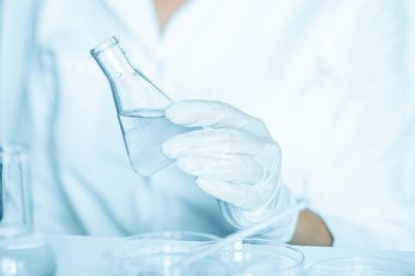 Laboratuvar içinde: yardımcı, laboratuvar ortamında görevleri yürütür. Laboratuvar önlüğü giyiyordu. Cam eşyalar kullanılıyor. Mavi laboratuvar içeriği. Şeffaf sıvıyla araştırma yapıyor..