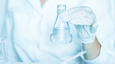 Laboratuvar aşaması: Asistan laboratuvar ortamında görevleri yerine getirir. Laboratuvar önlüğüyle süslenmiş. Cam teçhizatı kullanılıyor. Mavi bir laboratuarda bulundu. Şeffaf sıvıyla araştırmaya odaklanmış..