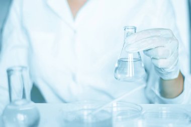 Laboratuvar kurulumu: Asistan laboratuvar koşullarında manipülasyon yapar. Laboratuvar önlüğü giyildi. Cam teçhizat kullanılmış. Mavi bir laboratuarda çalışıyor. Şeffaf sıvıyla araştırma yapıyor..