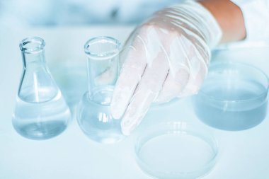 Laboratuvar içinde: yardımcı, laboratuvar ortamında görevleri yerine getirir. Laboratuvar önlüğü giyiyordu. Cam teçhizatı kullanılıyor. Mavi laboratuvar içeriği. Şeffaf sıvı kullanılarak yapılan araştırmalar.