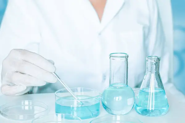 Laboratory scene: Lab assistant conducts procedures. Wearing gloves and a white robe. Employing glassware. Clear blue liquid.
