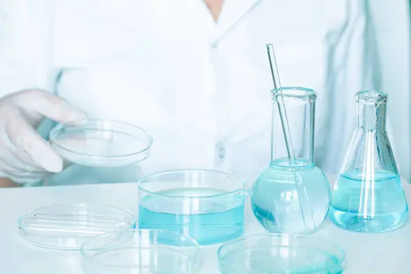 Lab work: Assistant performs actions in the laboratory. Wearing gloves and a white robe. Using glassware. Clear blue liquid present.