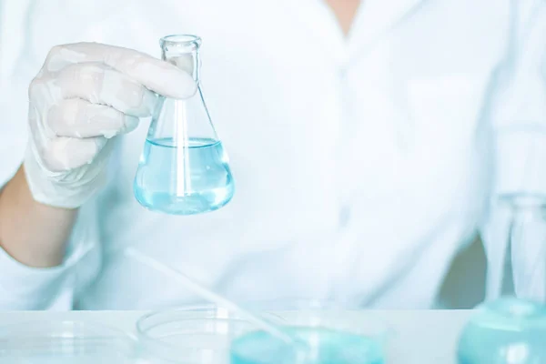Lab work: Aide conducts manipulations on flask\'s liquid. Gloves, white robe worn. Employing glassware. Clear blue liquid in focus.