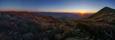 Arkasında günbatımı olan bir dağ silsilesinin panoramik görüntüsü. Gökyüzü turuncu ve pembe renklerin karışımıdır. Huzurlu ve huzurlu bir atmosfer yaratır.