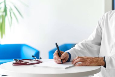 Hastanede steteskopla reçete yazan beyaz önlüklü bir doktor. Doktorlar beyaz bir arkaplan üzerinde elleri yakın çekim.
