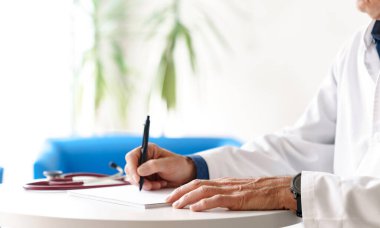 Hastanede steteskopla reçete yazan beyaz önlüklü bir doktor. Doktorlar beyaz bir arkaplan üzerinde elleri yakın çekim.