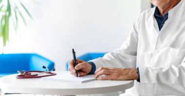 Hastanede steteskopla reçete yazan beyaz önlüklü bir doktor. Doktorun elleri beyaz arka planda..