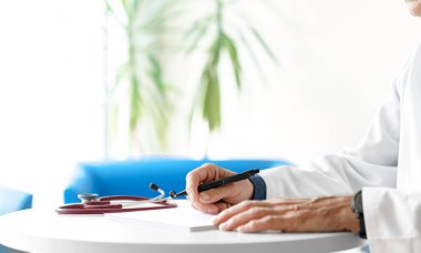 Hastanede steteskopla reçete yazan beyaz önlüklü bir doktor. Doktorun elleri beyaz arka planda..