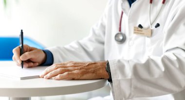 Hastanede steteskopla reçete yazan beyaz önlüklü bir doktor. Doktorun elleri beyaz arka planda..