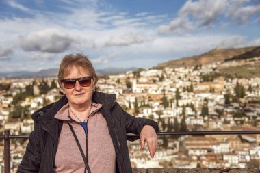 Güneş gözlüklü yaşlı kadın gülümsüyor ve Alhambra terasında kameraya bakıyor, arka planda Albaicin mahallesi var. Granada şehri, İspanya. Güneşli bir günde kışlık kıyafet giymenin tadını çıkarıyorum. Açık havada seyahat etmek..