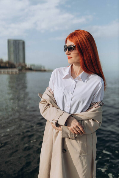 Stylish red hair lady stands, posing on the seaside in the afternoon, long brown trench-coat, sunglasses 
