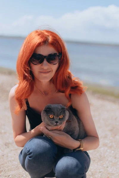 Red Hair lady holds beautiful Grey Scottish-fold shorthair fluffy cat with orange eyes outside near the river. Warm picture toning. Pets care. World domestic animal day. Image for websites about cats.